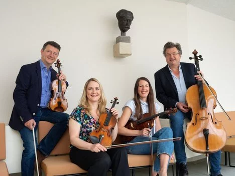 The image shows four smiling musicians with their string instruments: two violins, a viola, and a cello. They sit or stand relaxed in a bright room, with a bust hanging on the wall behind them.