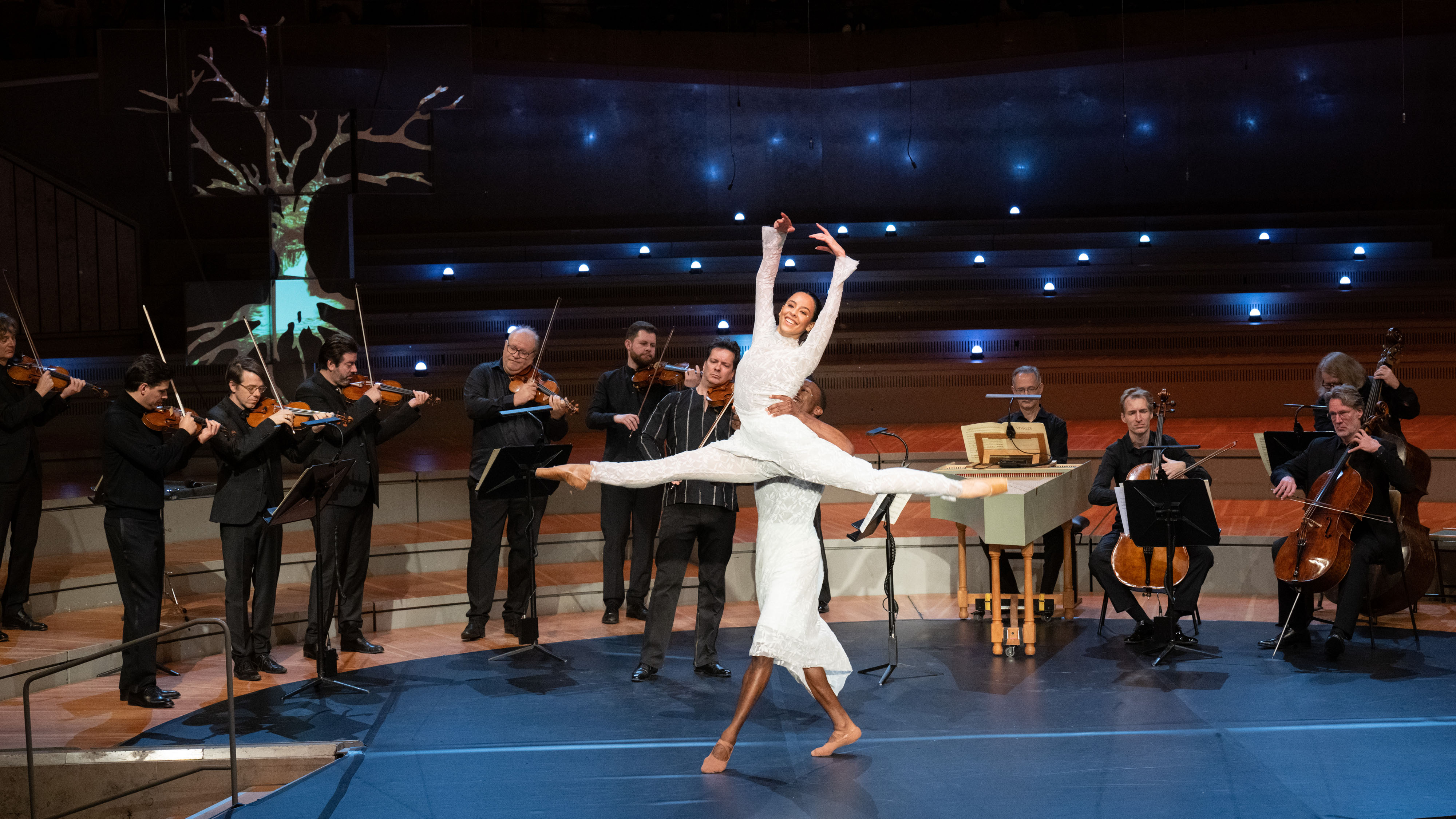 A dancer lifts a dancer, musicians play in the background