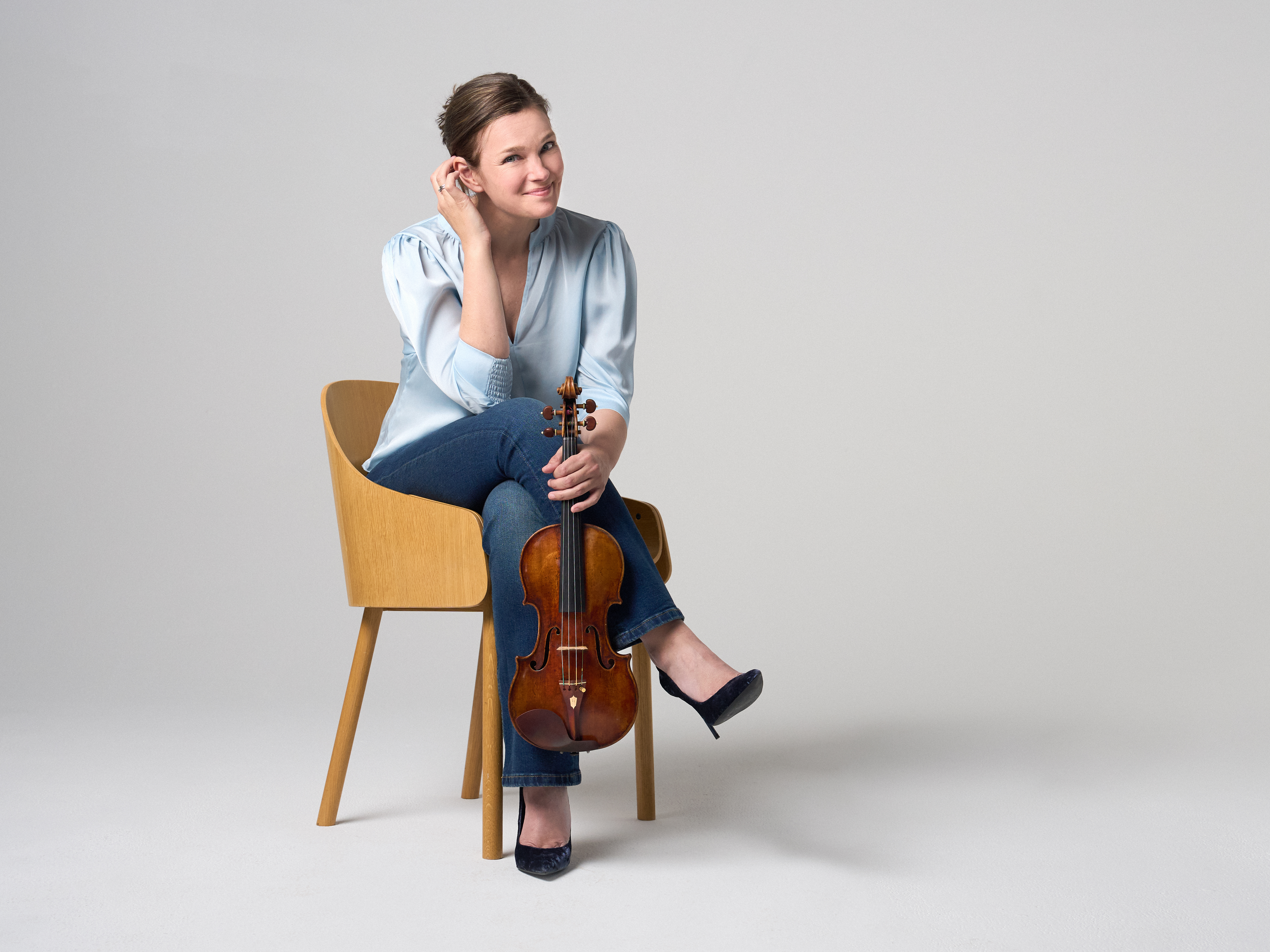 Janine Jansen is sitting on a chair, a violin in her hands. She is wearing jeans and a light blue blouse and looks into the camera with a smile.