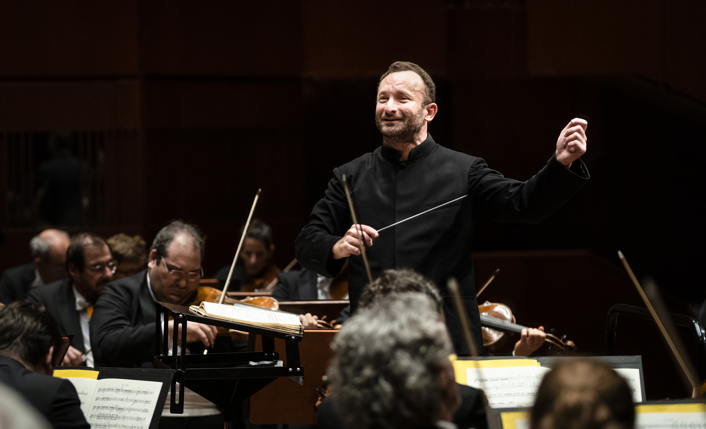Kirill Petrenko energetically conducts the orchestra. He holds a baton and gestures with his other hand, surrounded by musicians playing various string instruments.